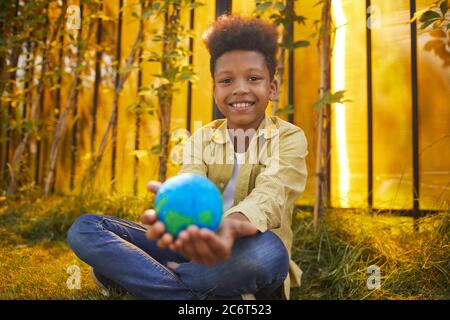 Portrait of smiling African-American boy holding planet in hands while sitting on green grass outdoors in sunlight, copy space Stock Photo
