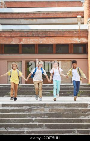 Vertical full length portrait of multi-ethnic group of children leaving school with backpacks and holding hands while running towards camera outdoors, Stock Photo