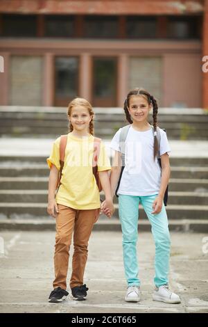 Vertical full length portrait of two cute schoolgirls with backpacks looking at camera while posing outdoors against school building Stock Photo