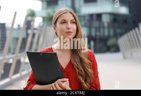 Serious business woman with folder documents standing among skyscrapers on modern city street and looking away Stock Photo