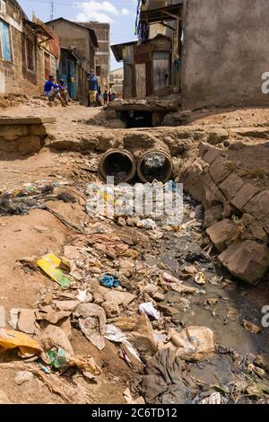 An open sewer filled with rubbish by the side of a dirt alley with residents nearby, Korogocho slum, Kenya, East Africa Stock Photo