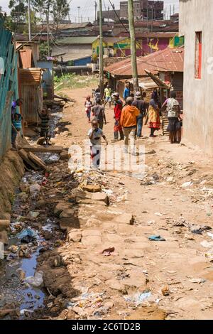 An open sewer filled with rubbish by the side of a dirt alley with residents nearby, Korogocho slum, Kenya, East Africa Stock Photo