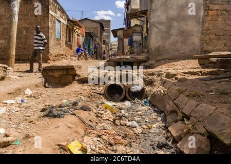 An open sewer filled with rubbish by the side of a dirt alley with residents nearby, Korogocho slum, Kenya, East Africa Stock Photo