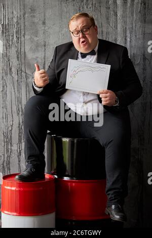 businessman happy with increasing of oil prices on global market, handsome guy sits on oil barrel, holding analysis chart showing current position Stock Photo