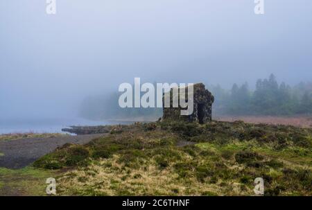 little stone hut covered by moss at the shallow banks of lake Lagoa Rasa surrounded by green forest lost in mist. mysterious moody day, the island of Stock Photo