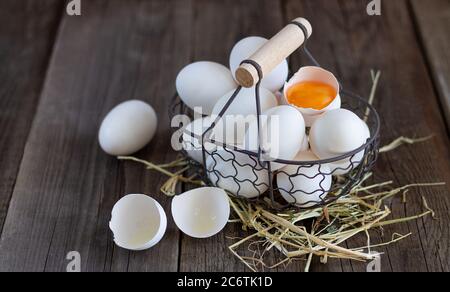 Fresh organic white eggs with eggshell and yolk in metal mesh basket on straw and wooden background. Country food concept Stock Photo