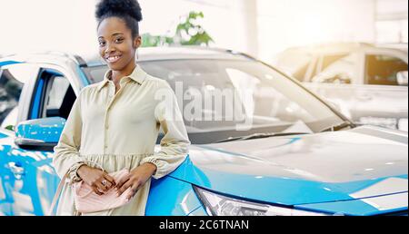 mixed race african american woman posing next to blue car in dealership, she wants to buy it Stock Photo