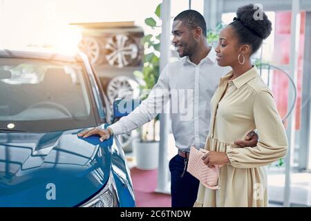 confident handsome afro man shows to wife a car he is liked, they look at car and discuss, man and woman think before making purchase Stock Photo