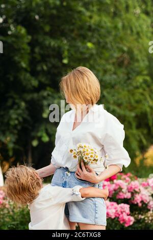 Unrecognizable blonde mother and son cuddling against the backdrop of the rosary. Stylish young family in white shirts and denim shorts. A bouquet of daisies. Stock Photo