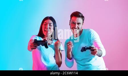 Virtual entertainment together. Happy guy and asian girl with joysticks in hands, rejoices to victory Stock Photo