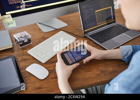 High angle closeup of female hands holding smartphone with code on screen while working at desk in office, female IT developer concept, copy space Stock Photo