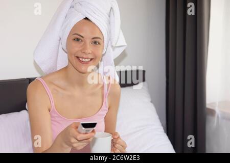 A young woman is holding a TV remote control while sitting on a bed in a hotel room. Stock Photo