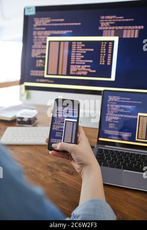 Vertical close up of female hand holding smartphone with code on screen while working at desk in office, female IT developer concept, copy space Stock Photo