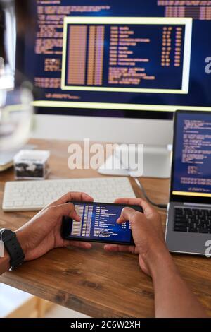 Vertical closeup of male hands holding smartphone with code on screen while working at desk in office, IT developer concept, copy space Stock Photo