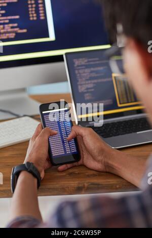 Unrecognizable African-American man holding smartphone with code on screen while working at desk in office, IT developer concept, copy space Stock Photo