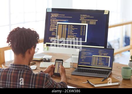 Back view portrait of modern African-American man holding smartphone with code on screen while working at desk in office, IT developer concept, copy s Stock Photo