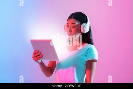 Shock in social network. Surprised asian girl in big headphones looks at tablet with luminous screen Stock Photo