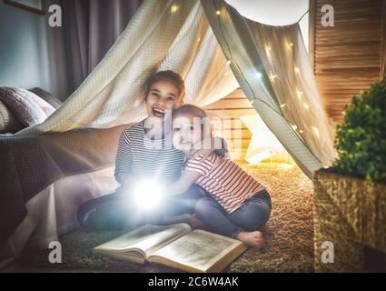 Two cute little children are reading a book with flashlights in tent. Happy girls playing at home. Funny lovely kids having fun in room. Stock Photo