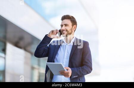 Busy young entrepreneur communicating on cellphone and looking through document on tablet computer at city Stock Photo