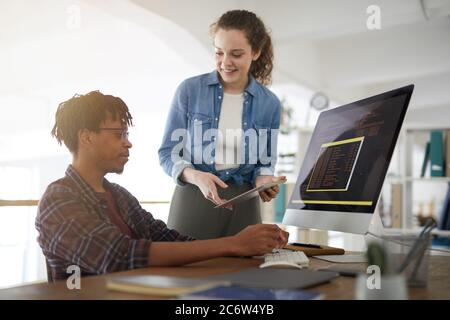 Portrait of smiling young woman talking to African-American man writing code while working in software development agency, copy space Stock Photo