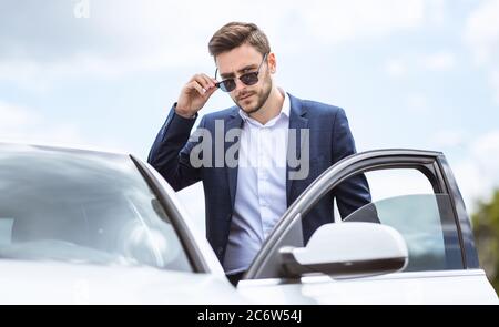 Serious CEO in formal wear and sunglasses getting into his automobile at downtown area of modern city Stock Photo