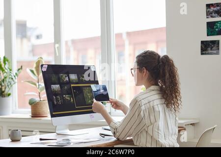 Back view portrait of modern young woman holding photographs reviewing for publishing while working at PC in white office, copy space Stock Photo