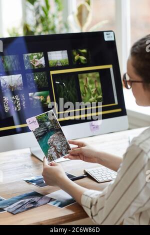 Vertical portrait of young female photographer holding pictures and smiling while sitting at desk with photo editing software on computer screen Stock Photo