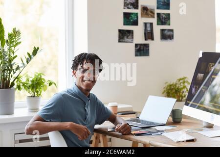 Portrait of young African-American photographer smiling at camera while posing at desk with photo editing software on computer screen, copy space Stock Photo