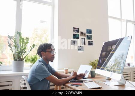 Portrait of young African-American photographer using computer at desk in home office with photo editing software on screen, copy space