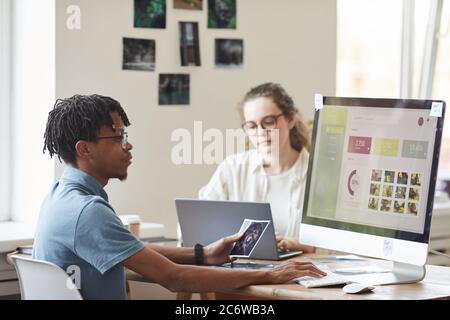 Portrait of young African-American photographer managing photo stock marketplace while using computer at desk in office with website on screen, copy s Stock Photo