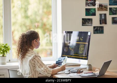 Side view portrait of young female photographer wearing mask while using computer at desk in homeoffice with photo editing software on screen, copy sp Stock Photo