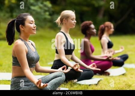 Concentrated multinational ladies meditating or doing breathing exercises on yoga practice outside, empty space Stock Photo