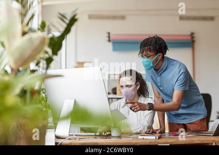 Portrait of two young photographers wearing masks while pointing at computer screen and reviewing pictures in office studio, copy space Stock Photo