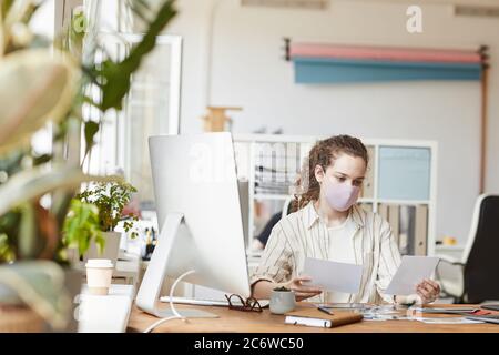 Front view portrait of young female photographer wearing mask while reviewing pictures sitting at desk in home studio, copy space Stock Photo