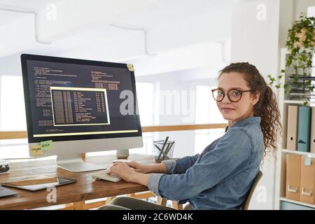 Portrait of young woman looking at camera while writing programming code on computer screen and working at desk in contemporary office interior, copy Stock Photo