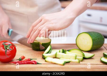 Female hands cut with a knife a young seasonal zucchini into strips on a wooden cutting board. The method of preparation of vegetables. Seasonal produ Stock Photo