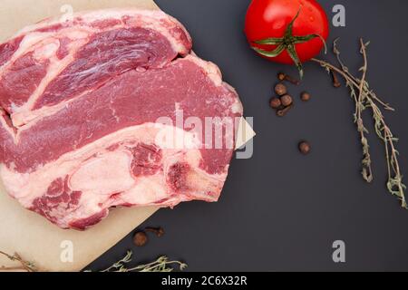 Fresh raw beef steak with spices, herbs and vegetables. The meat is on the bone. Thyme, black pepper, tomatoes, beef on black stone. Food background. Stock Photo