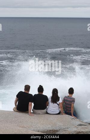 A group of four visitors sit very close to crashing waves on the rocks at Peggy’s Cove, Nova Scotia, Canada despite warnings about the danger of the A Stock Photo