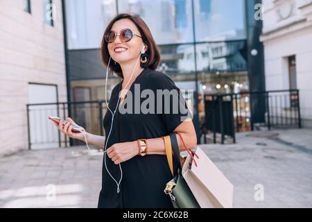 Smiling beautiful modern middle-aged female Portrait dressed black dress and sunglasses with shopping bags holding slim smartphone listening music wit Stock Photo