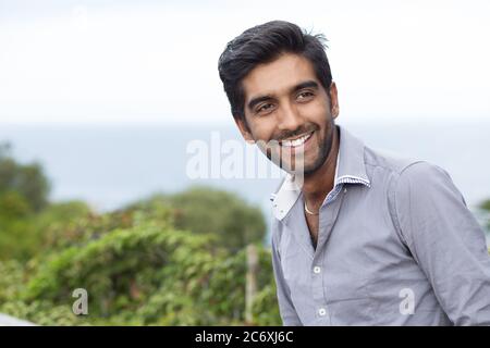 Smiling happily man with cheerful face expression standing outdoors in the street, green bushes and sea on background, toothy smile middle age man mul Stock Photo