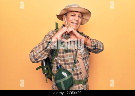 Senior man with grey hair wearing hiker backpack and water canteen smiling in love doing heart symbol shape with hands. romantic concept. Stock Photo