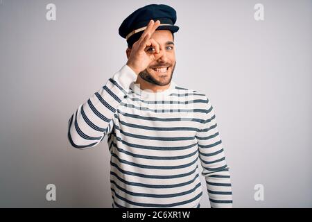 Young handsome sailor man with beard wearing navy striped uniform and captain hat doing ok gesture with hand smiling, eye looking through fingers with Stock Photo