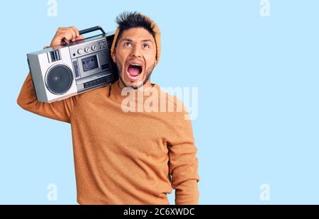 Handsome latin american young man holding boombox, listening to music angry and mad screaming frustrated and furious, shouting with anger. rage and ag Stock Photo
