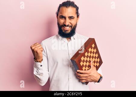 Young arab man holding chess screaming proud, celebrating victory and success very excited with raised arm Stock Photo