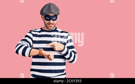 Young handsome man wearing burglar mask in hurry pointing to watch time, impatience, upset and angry for deadline delay Stock Photo