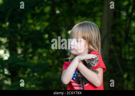 A five-year-old girl holding a tabby kitten outdoors in summer with trees in background. Stock Photo