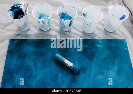 arts and craft hobbies concept, blue and black acrylic painting with colors in plastic cups and mixed painting tools next to it while the work is in p Stock Photo