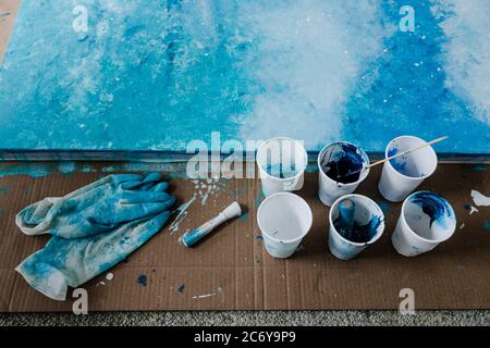 arts and craft hobbies concept, blue and black acrylic painting with colors  in plastic cups and mixed painting tools next to it while the work is in p  Stock Photo - Alamy