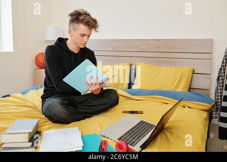 Software engineering student sitting on bed with books, laptop and textbooks around and writing in notebook Stock Photo