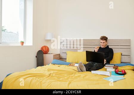 Positive high school student sitting on bed, eating pizza and watching online webinar when staying home during quarantine Stock Photo